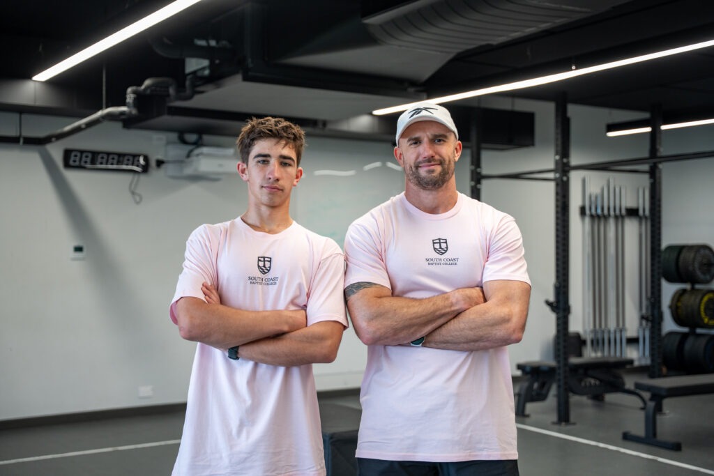 Medium portrait of Coach Andy Price and an SCBC student athlete smiling slightly with arms crossed, wearing matching pastel pink South Coast Baptist College sports shirts in the gym.