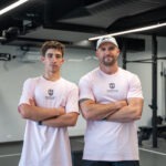 Medium portrait of Coach Andy Price and an SCBC student athlete smiling slightly with arms crossed, wearing matching pastel pink South Coast Baptist College sports shirts in the gym.